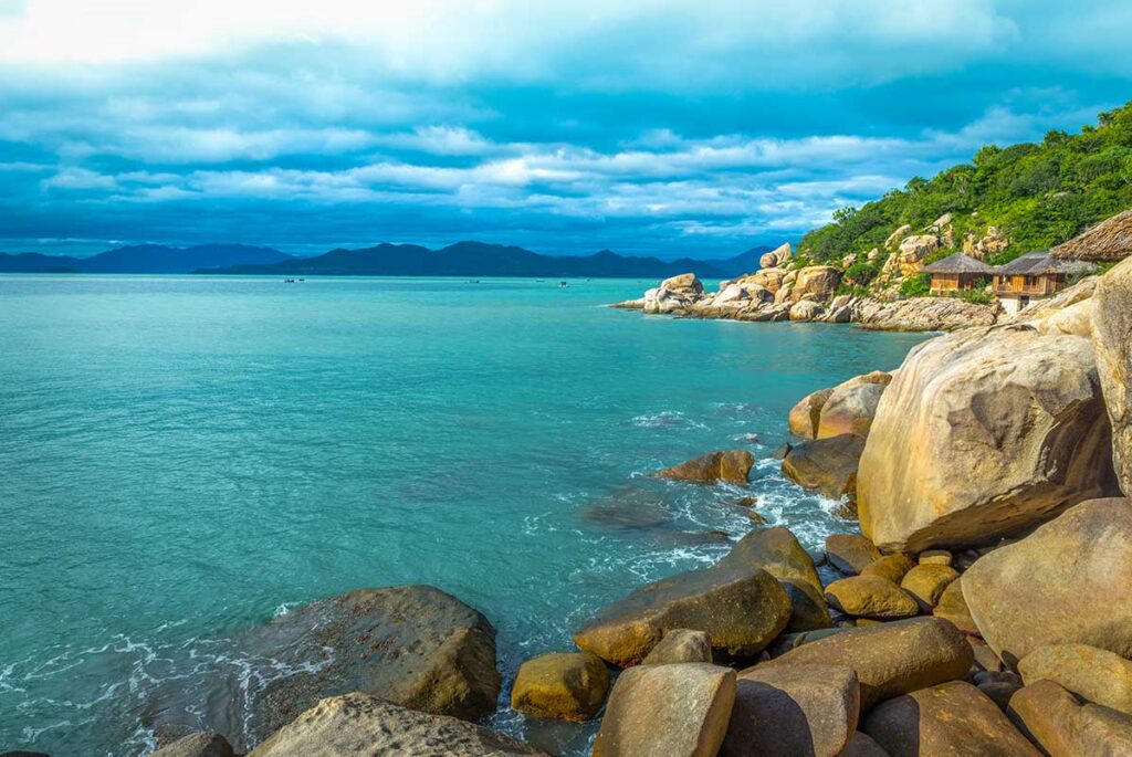 Felsige Küstenlinie der Ninh Van Bay mit Granitfelsen, türkisfarbenem Wasser und Reetbungalows eines Luxusresorts versteckt zwischen den bewaldeten Hügeln im Hintergrund