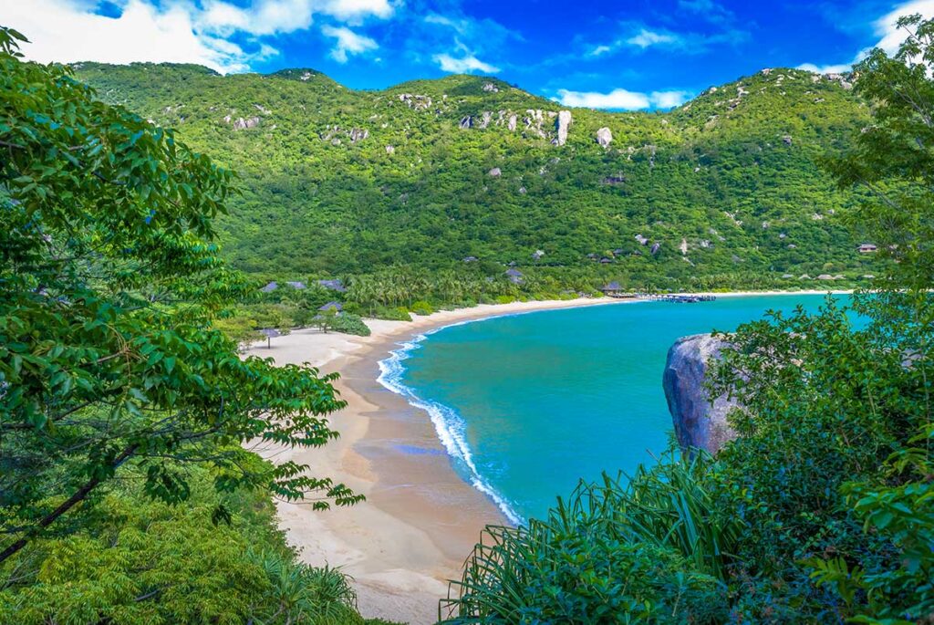 Blick aus dem Dschungel auf die Bucht von Ninh Van mit einem langen weißen Strand, türkisfarbenem Wasser und Reetbungalows eines Resorts, das in die grünen Berghänge integriert ist