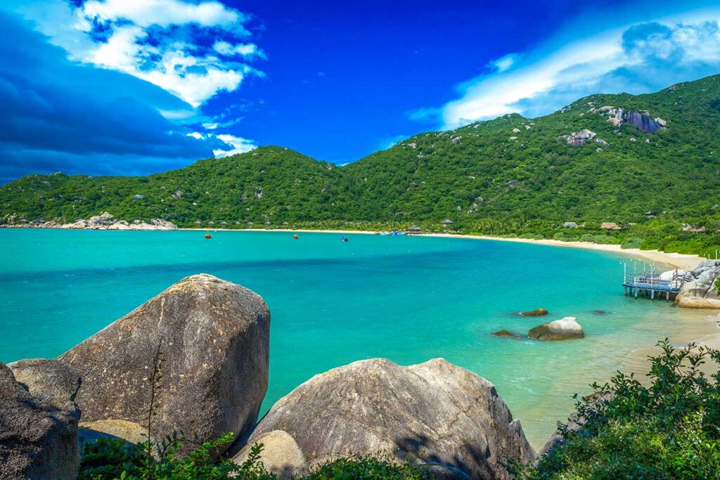 Panoramablick auf die Ninh Van Bay mit Granitfelsen im Vordergrund, türkisfarbenem Wasser, einem weißen Sandstrand und Reetbungalows eines Luxusresorts entlang der Küstenlinie