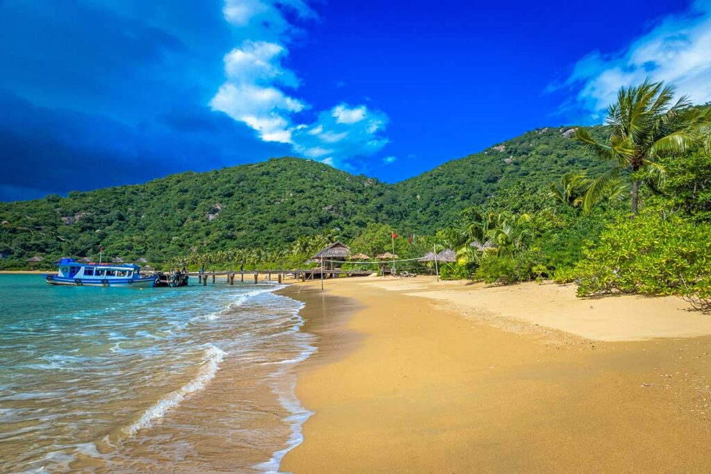 Breiter goldgelber Strand der Ninh Van Bay mit weißen Wellen, Palmen und einem Holzsteg in der Ferne — der Strand ist Teil eines exklusiven Resorts, das nicht öffentlich zugänglich ist