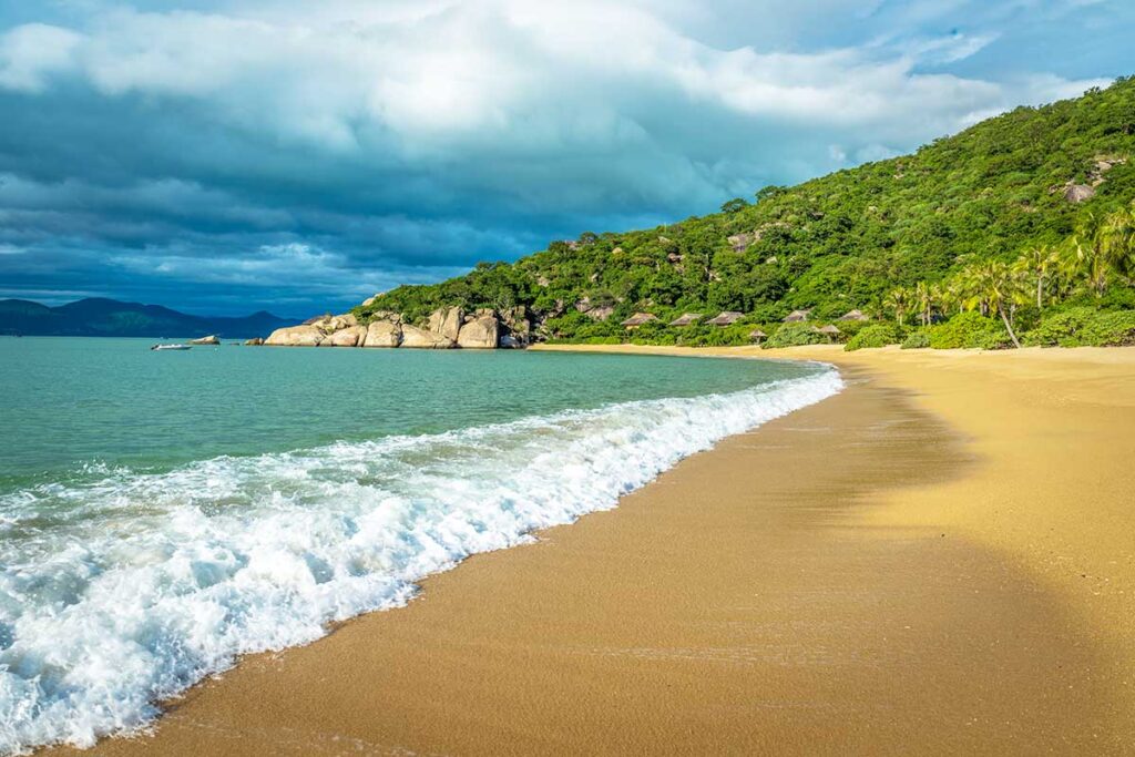 Breiter goldgelber Strand der Ninh Van Bay mit anrollenden Wellen, Palmen und Reetbungalows im Dschungel — eine der schönsten und abgelegensten Buchten bei Nha Trang