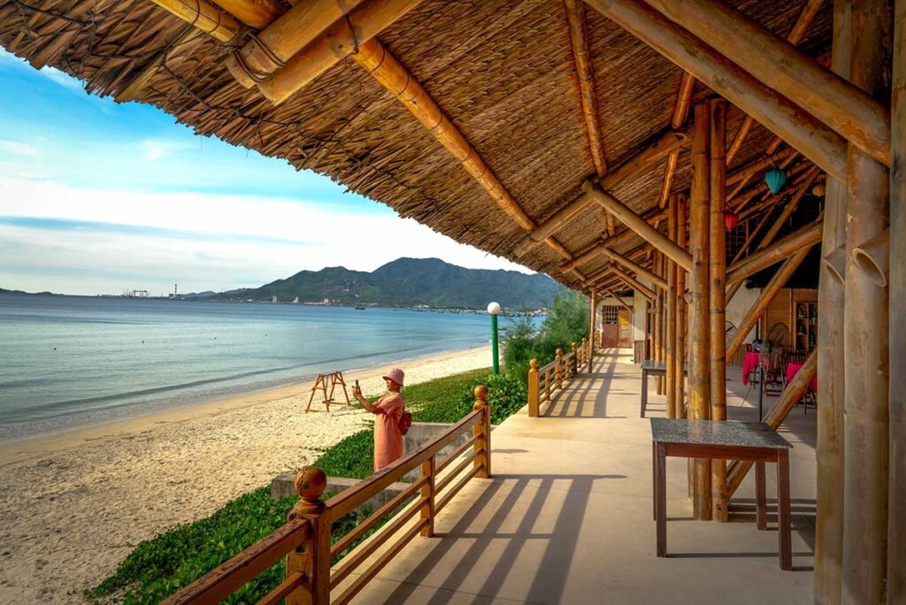 Überdachte Terrasse eines Strandresorts bei Doc Let Beach mit Bambuspfeilern und Reetdach, Blick auf das ruhige Meer und einer Besucherin, die ein Foto auf dem weißen Sand macht