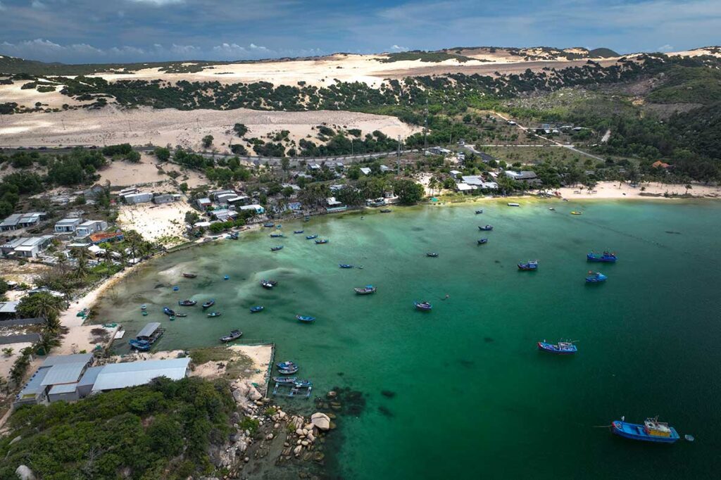 Luftaufnahme des Fischerhafens von Dam Mon mit Dutzenden bunter Boote auf dem grünen Wasser, einem kleinen Fischerdorf und Sanddünen im Hintergrund in der Van Phong Bay
