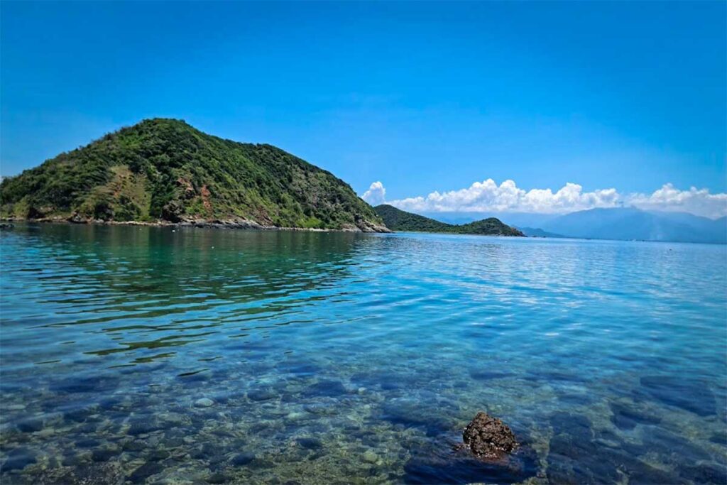 Kristallklares Wasser in der Van Phong Bay mit sichtbaren Steinen auf dem Grund und einem bewaldeten Hügel im Hintergrund — das klare Wasser macht das Schnorcheln in der Van Phong Bay sehr attraktiv