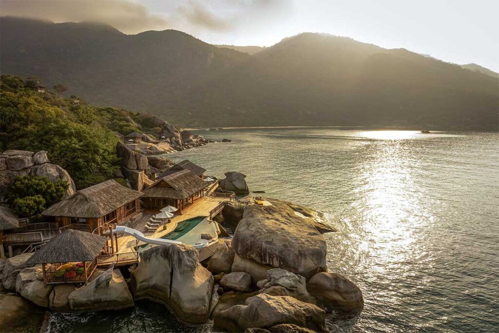 Luftaufnahme des Six Senses Resorts entlang der felsigen Küstenlinie der Ninh Van Bay im Abendlicht, mit Infinity-Pool, Holzterrassen und Bungalows verteilt über die Felsen am Wasser