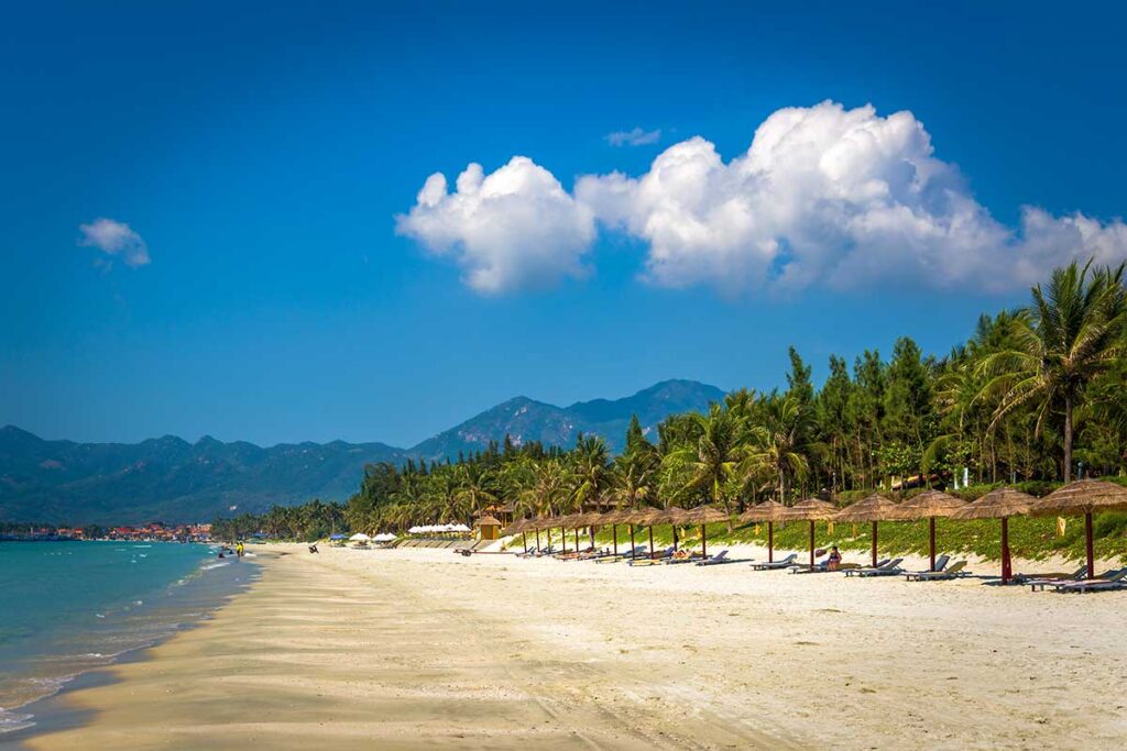 Langer weißer Strand von Doc Let Beach mit Reetschirmen, Liegestühlen, Palmen und Bergen im Hintergrund — der Strand liegt etwa eine Stunde von Nha Trang entfernt und zieht verhältnismäßig wenige Touristen an