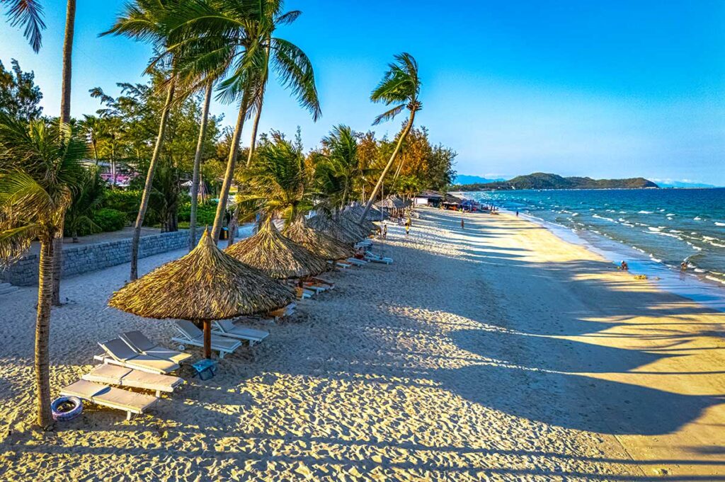 Reihe von Reetschirmen und Liegestühlen am weißen Strand von Doc Let Beach im Abendlicht, mit im Wind bewegenden Palmen und dem Meer im Hintergrund