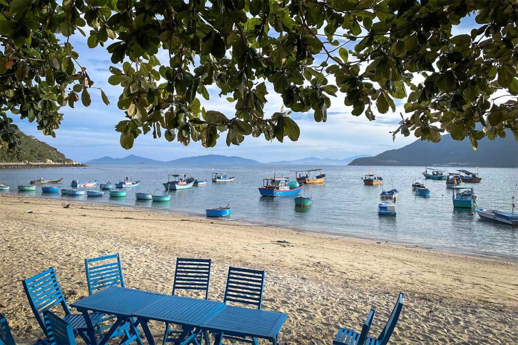 Strand des Fischerdorfs Ninh Van mit blauen Stühlen und Tischen unter einem Baum, Dutzenden runder und hölzerner Fischerboote im flachen Wasser und Bergen am Horizont