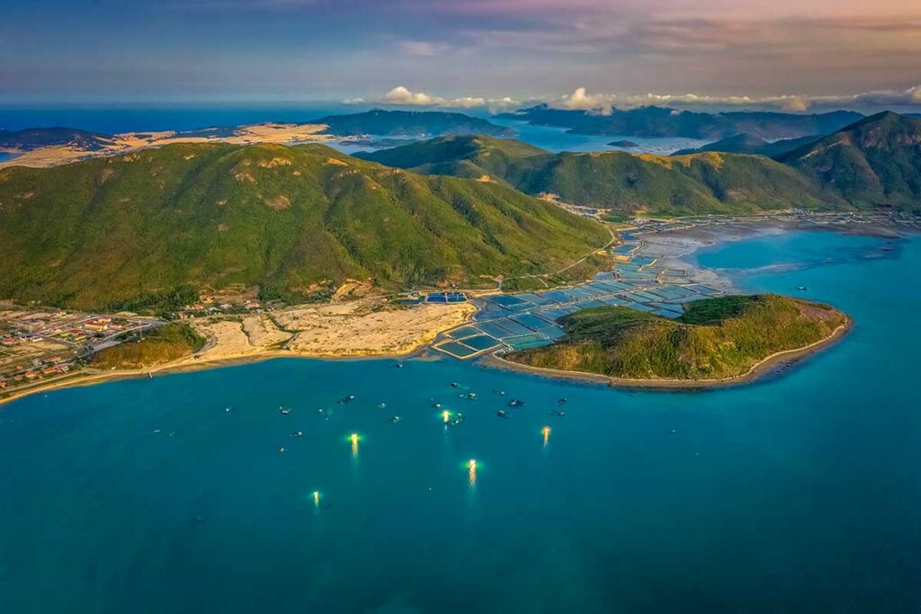 Luftaufnahme der Van Phong Bay im Abendlicht mit grünen Berghängen, Fischzuchtanlagen an der Küste und Fischerbooten mit gelben Lichtern auf dem türkisfarbenen Wasser