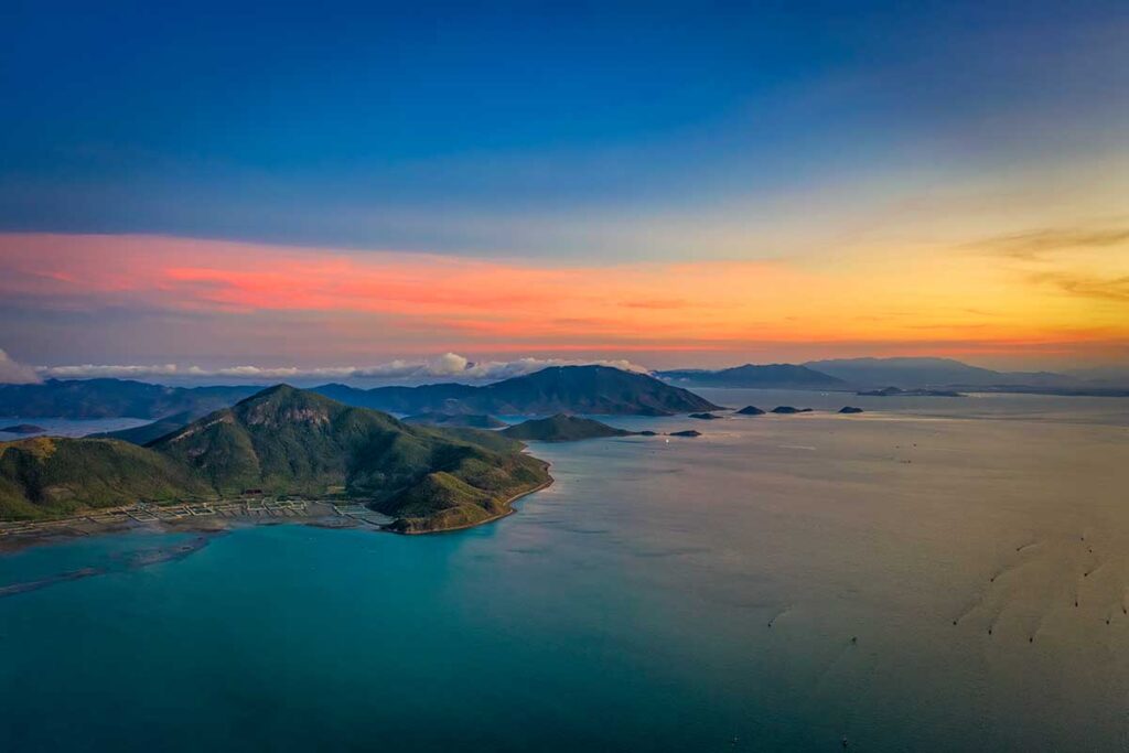 Luftaufnahme der Van Phong Bay bei Sonnenuntergang mit einem farbenfrohen Himmel in Rosa-, Orange- und Blautönen, grünen Berginseln und ruhigem Wasser — eine der schönsten Buchten nördlich von Nha Trang