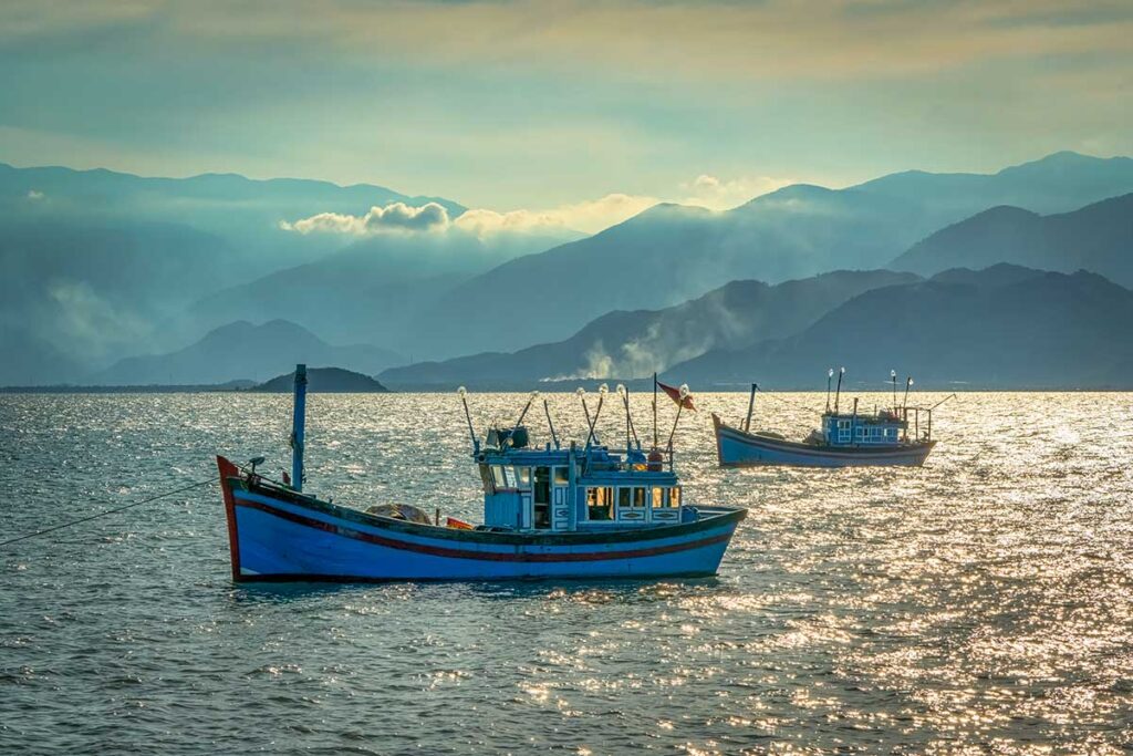 Zwei traditionelle vietnamesische Fischerboote auf dem glitzernden Wasser der Van Phong Bay mit Bergketten im Dunst im Hintergrund — ein authentisches Bild des Fischerlebens in der Bucht