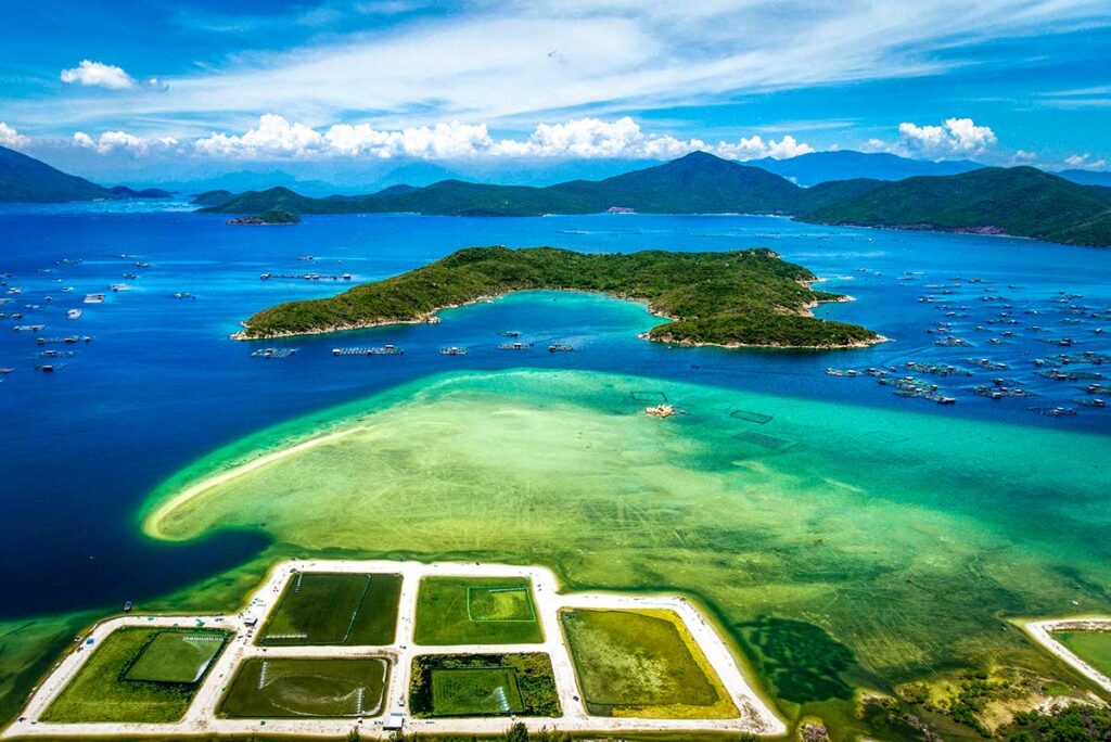 Luftaufnahme der Van Phong Bay mit Hon Lon Insel im Zentrum, umgeben von türkisem und tiefblauem Wasser, Fischzuchtanlagen und mehreren kleineren Inseln verteilt über die Bucht